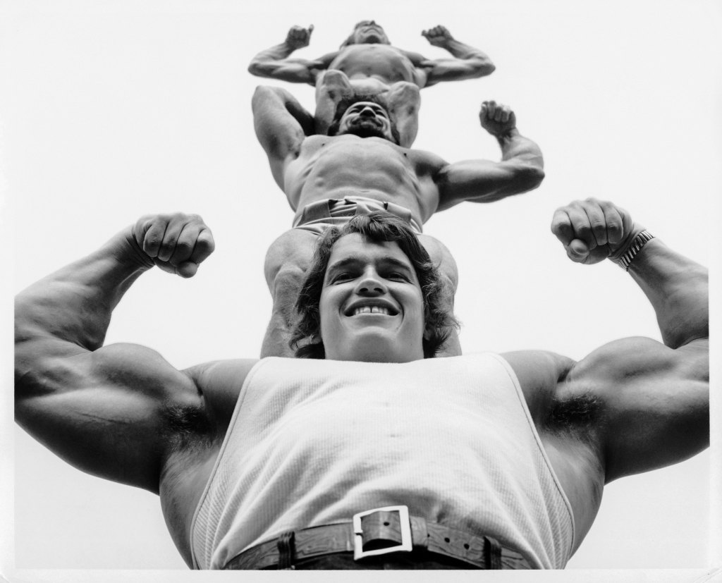 black and white image of Arnold Schwarzenegger holding two man on his shoulders as a human tower