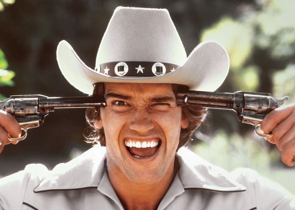 Arnold Schwarzenegger wearing a cowboy hat holding two pistols up to his head