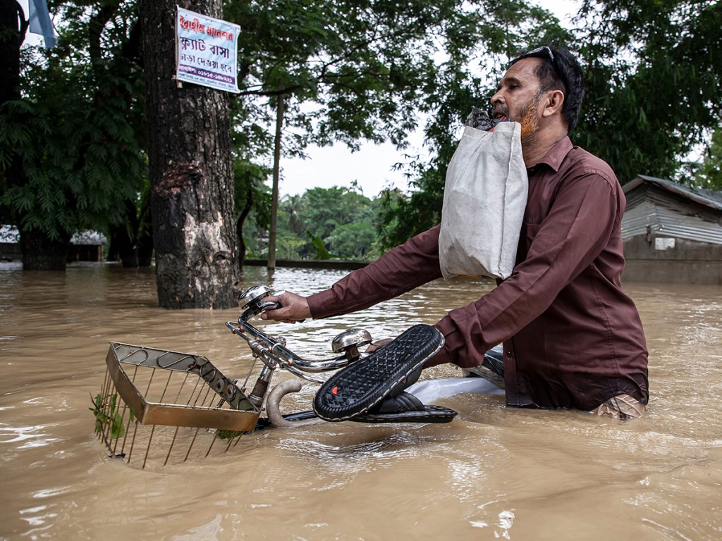 A man carries a bicycle and emergency supplies while wading through waist-deep floodwater in Satkania, Chattogram district, Bangladesh,