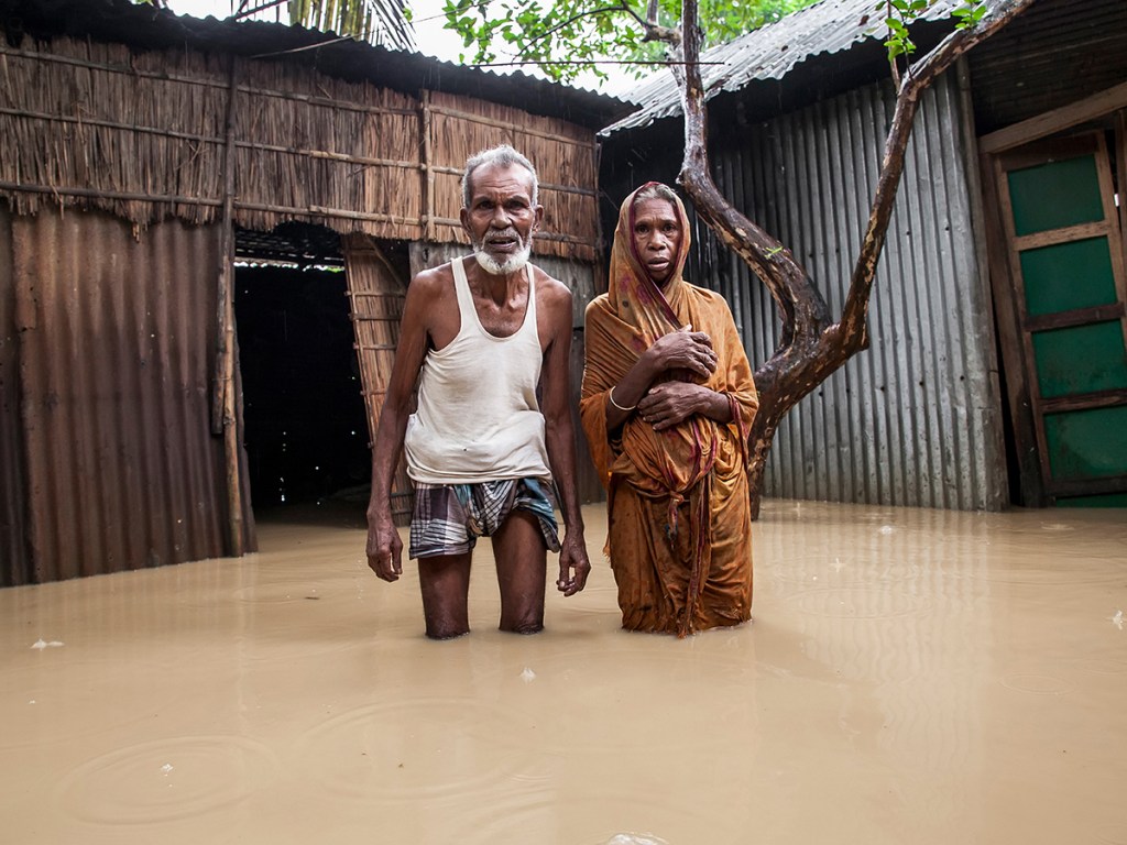 An elderly couple stands in front of their home surrounded by knee-deep floodwater in Jhenaigati upazila, 