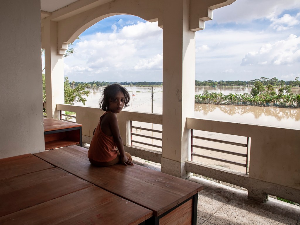 A child sits on a table inside a flood shelter while looking toward the flooded landscape outside in Chhagalnaiya upazila, Feni district, Bangladesh