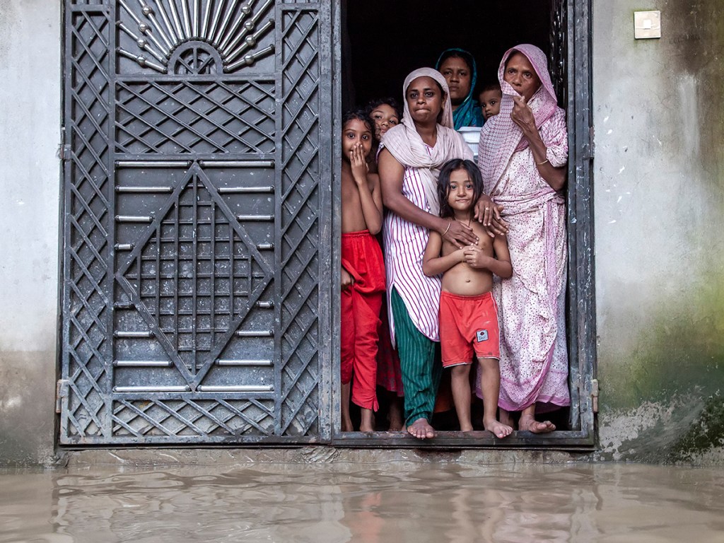 Members of a family stand at the entrance of their flooded house in Mirsharai upazila, Chattogram
