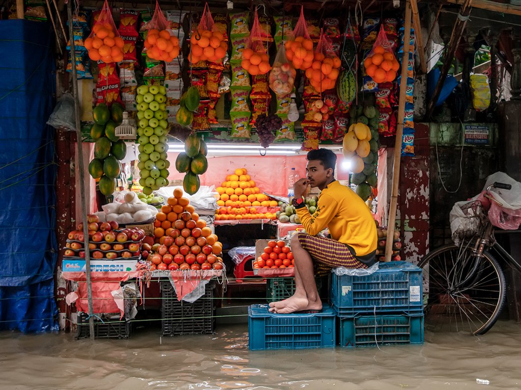 A fruit vendor waits for customers while sitting on stacked plastic crates to avoid floodwater in Chattogram city,