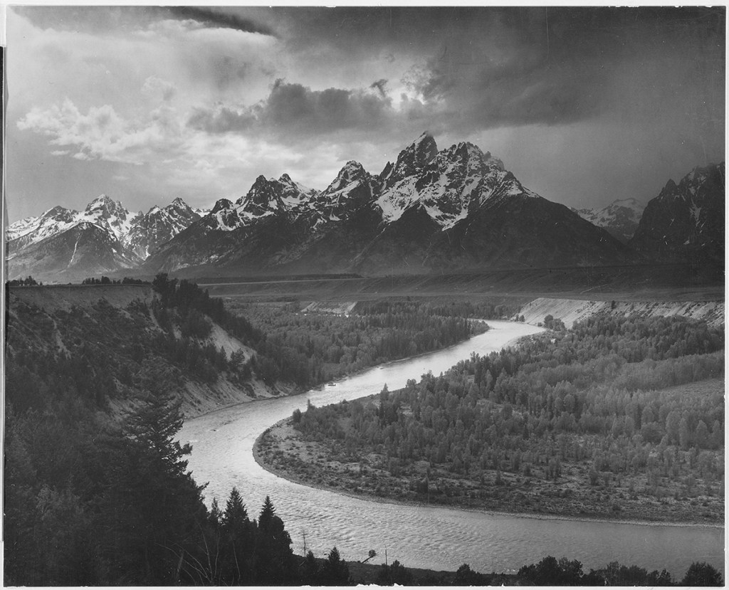 A black and white photograph of several of the peaks of the Teton Range covered in snow, the Snake River winds through the landscape and is surrounded by heavily forested lands; the image comes from a series of photographs known as the mural project by Ansel Adams, who was commissioned by the National Park Service in 1941 to create a photographic mural for the Department of the Interior Building in Washington DC. The project came to an end due to World War II. Wyoming, United States, 1941. (Photo by Smith Collection/Gado/Getty Images).
