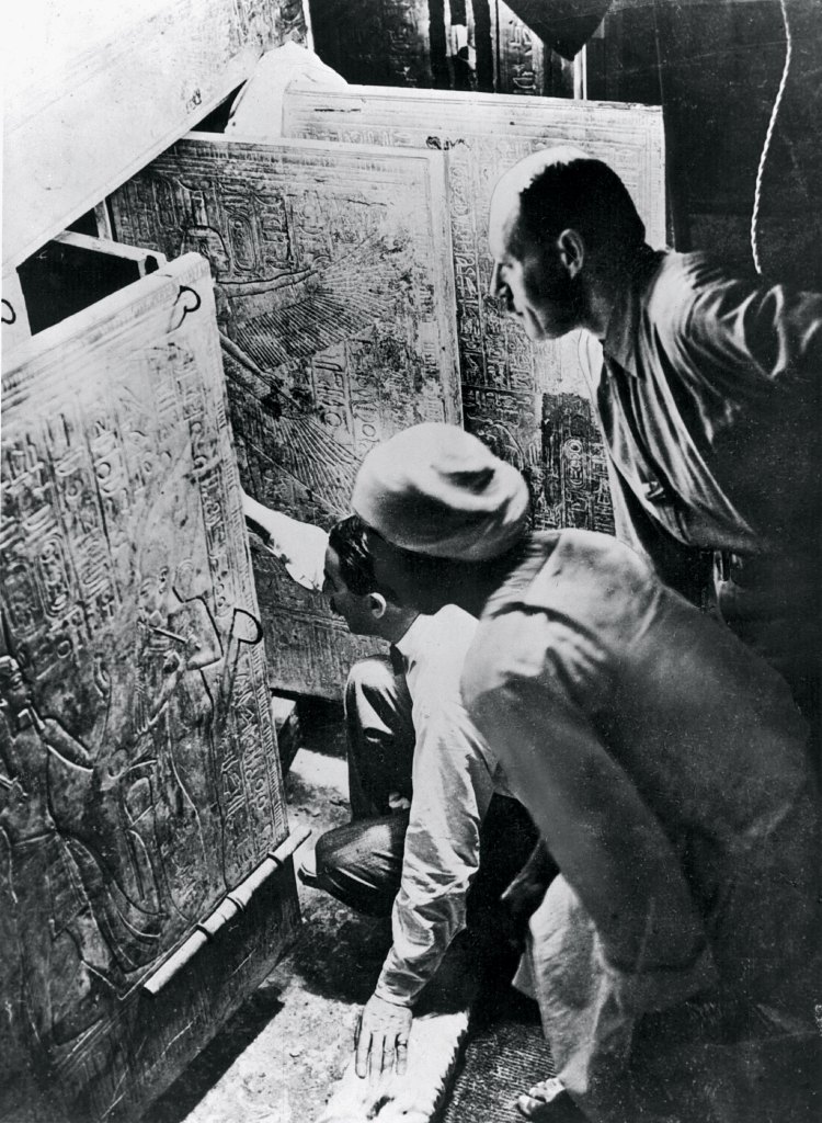 A historic black and white photograph by Harry Burton showing Howard Carter looking through the open gilded shrines of King Tut's tomb.