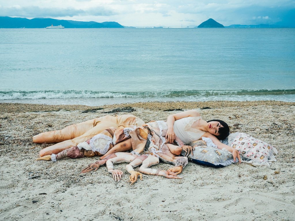 Colour photograph of a woman lying on her side on a beach. Spread out around her are a number of fake limbs.