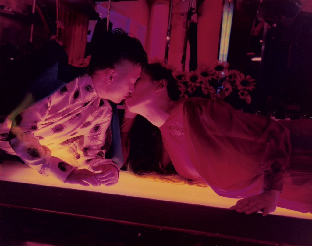 Colour photograph of two people leaning over to kiss each other.