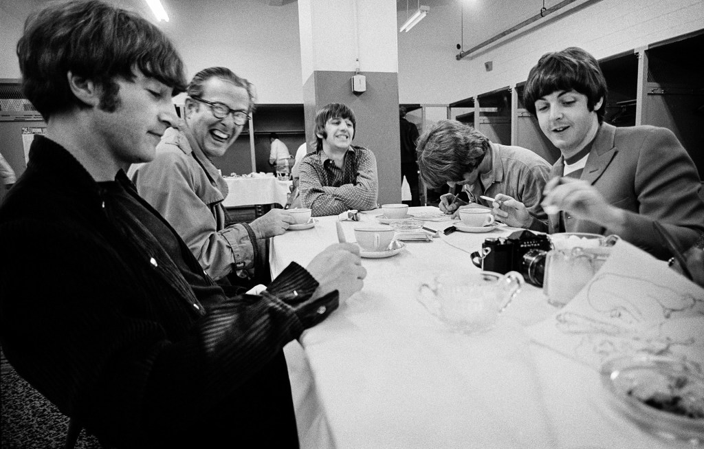John Lennon, Ralph J. Gleason, Ringo Starr,
George Harrison, Paul McCartney, and Joan Baez doodle on
the tablecloth in the locker room at Candlestick Park. Simpson’s Catering sets up in the background. Image Credit: 2026 © Jim Marshall ONE TIME USE