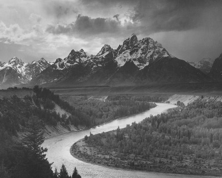 A black and white photograph of several of the peaks of the Teton Range covered in snow, the Snake River winds through the landscape and is surrounded by heavily forested lands; the image comes from a series of photographs known as the mural project by Ansel Adams, who was commissioned by the National Park Service in 1941 to create a photographic mural for the Department of the Interior Building in Washington DC. The project came to an end due to World War II. Wyoming, United States, 1941. (Photo by Smith Collection/Gado/Getty Images).
