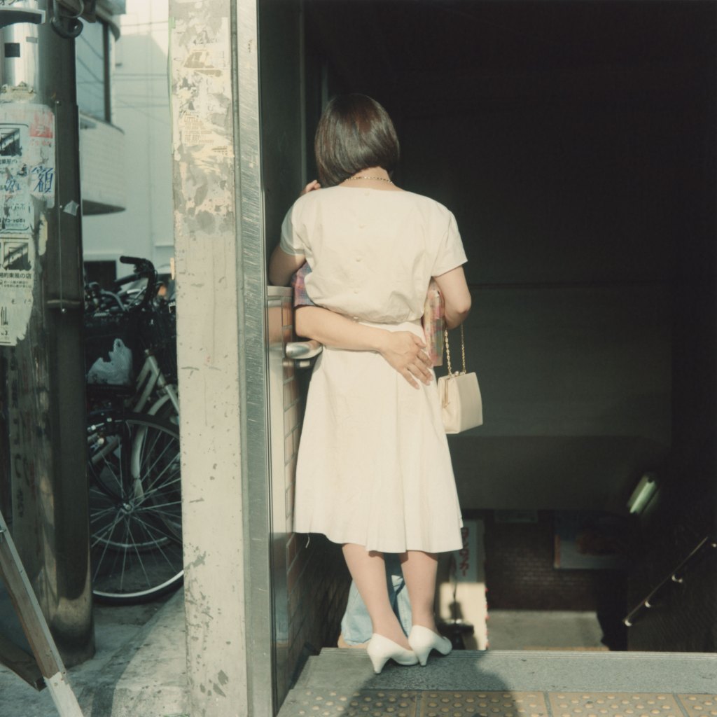 Colour photograph of the back of a woman standing against a wall. An arm can be seen around her waist.