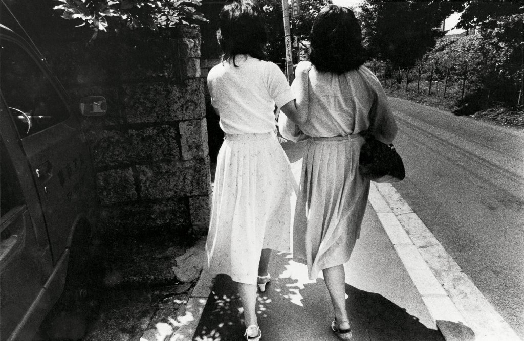 Black and white photograph of two women walking along a pavement.