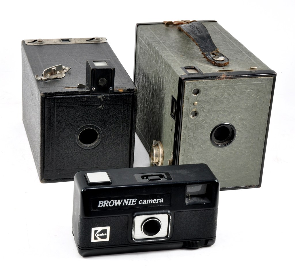 In 1900, the first camera to bear the Brownie name was aimed at children, at a time when brownies in America were pixies or elves. The camera had no viewfinder, only two V-shaped lines etched into the top to line up on the subject, although a separate accessory viewfinder, seen attached here, was available. The first Brownie (second version) is pictured here with a green No.2 Brownie, the first camera to use 120 size roll film and a Brownie 110 from 1980, the last camera to bear the Brownie name