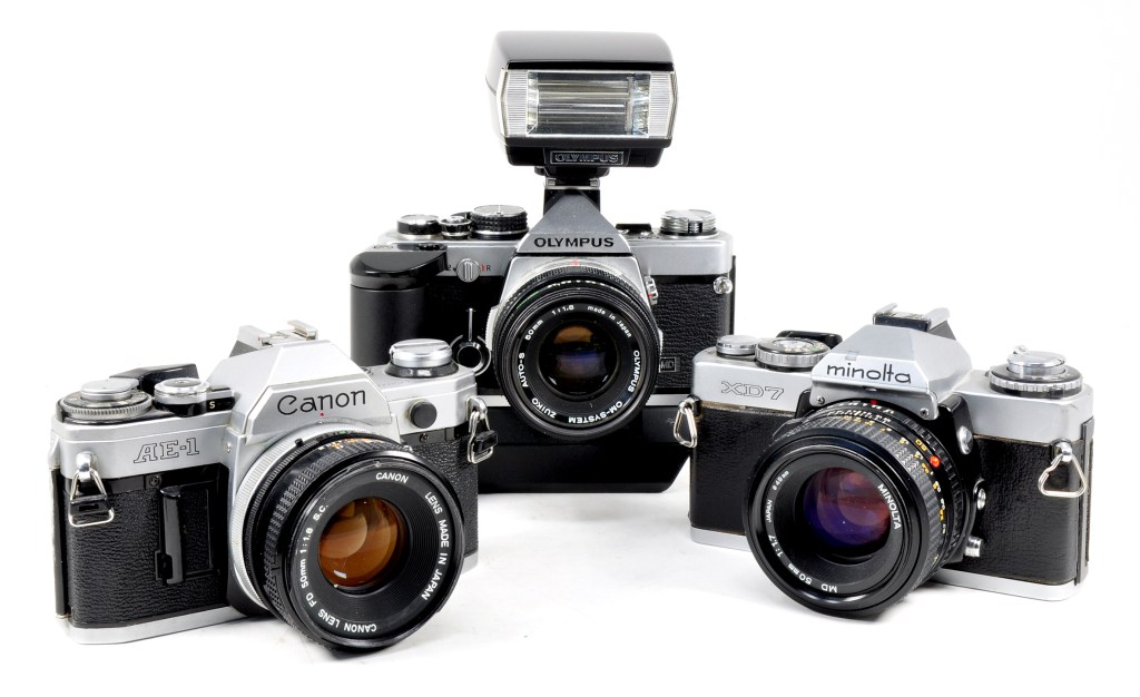 Metering pioneers: Olympus OM-2 illustrated with T20 flashgun and OM Winder 2 fitted, Canon AE-1 and Minolta XD-7