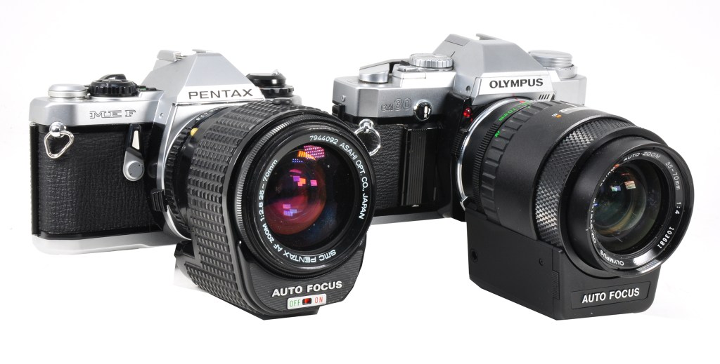 Early autofocus relied on special lenses for attachment to manual focus SLRs. Here are the Pentax ME F, the first camera specifically made as an autofocus SLR, and the Olympus OM30, with its purpose-made autofocus lens