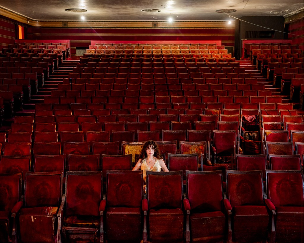 A woman sits alone in the middle of a large empty theatre with rows of red chairs