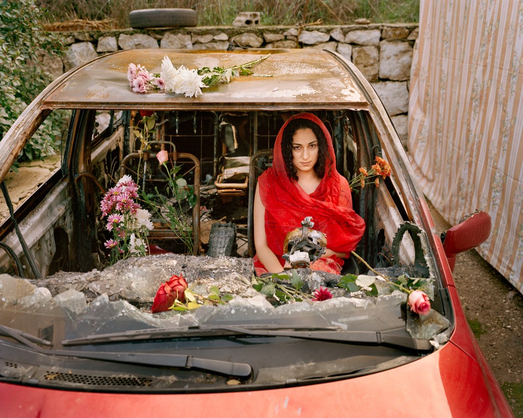 A woman wearing a red dress and headscarf sitting in a wrecked car decorated with flowers