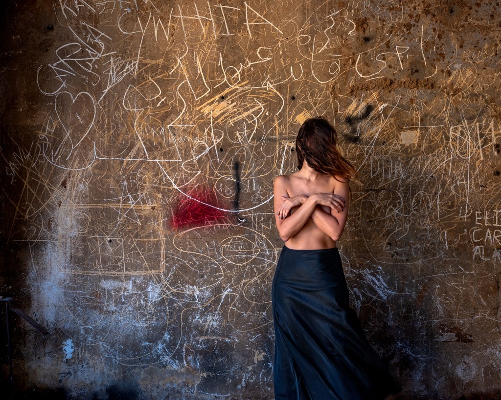 A woman standing against a wall full of inscriptions, her upper body only covered by her hands while her face is obscured by her hair