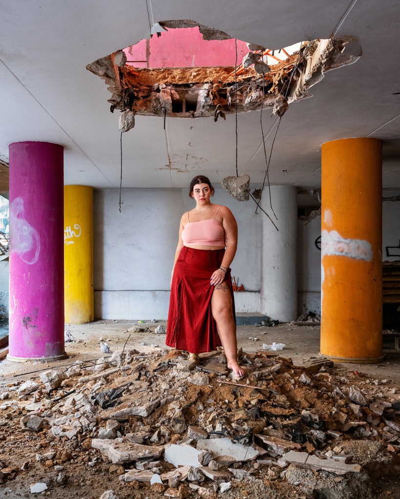 A women wearing a red skirt and pink blouse stands on top of rubble inside a derelict building with colourful pillars