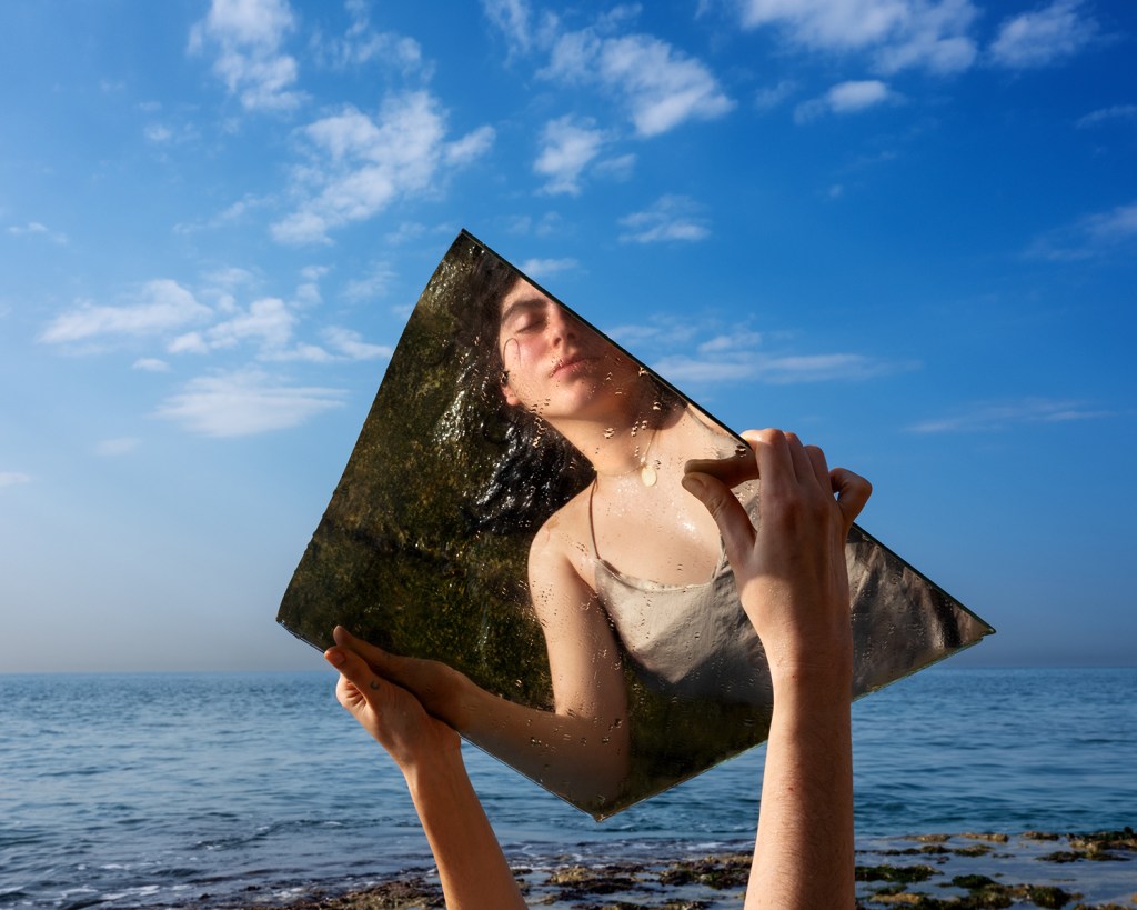 A woman's reflection photographed as she is holding up a mirror with the sea in the background