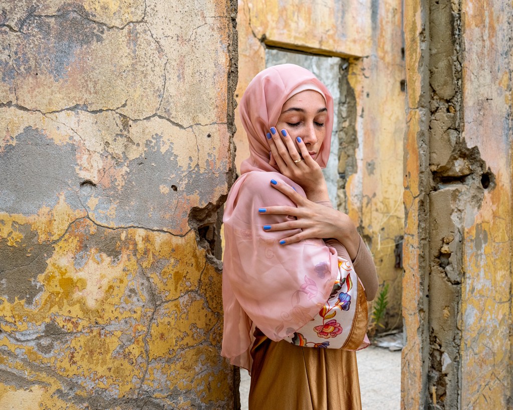 A woman wearing a pink headscarf poses with her shoulder turned towards the camera, her hand half covering her face
