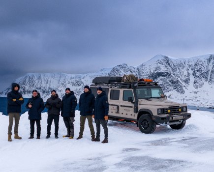 six man stand for a photo next to a touring car against a snowy landscape and icy mountain in the background