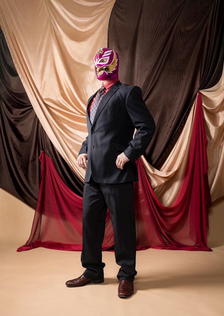 Capturing a heroic view of an icon, this photograph presents a Mexican wrestler reimagined as a sharp, elegant businessman. This represents all the hard work behind the theatrical sport which many enjoy, holding tradition and originality. Framed by stage curtains, the portrait highlights the rich comedy and dramatic artistry behind "lucha libre."