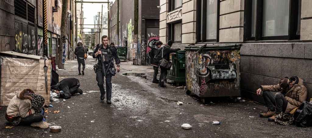A heavily armed Vancouver policeman patrols an alley surrounded by drug users outside a public overdose prevention site off East Hastings after a report of threatening behaviour. Image Credit © Simon Townsley