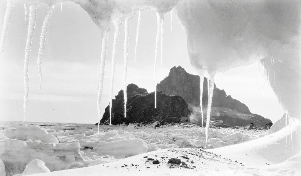 Hanging icicles stalactites frame the opening of an ice cave. We are looking out into the open with an iceberg or mountain in the background