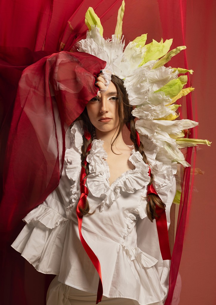 'La Bandera'. Green, white, and red: the living canvas of the Mexican flag. In this image, green evokes the hope carried in our hands, white mirrors the purity of our people's hearts, and red honours the blood spilled by those who fought for our freedom. Wrapped in the depth of Mexican history, the model stands with unmistakable pride, holding fast to the red. Her headpiece features corn leaves, a sacred tribute to the ancient symbol of life. Together, these elements transform the photograph into a profound visual love letter to our country, born straight from the flag itself.