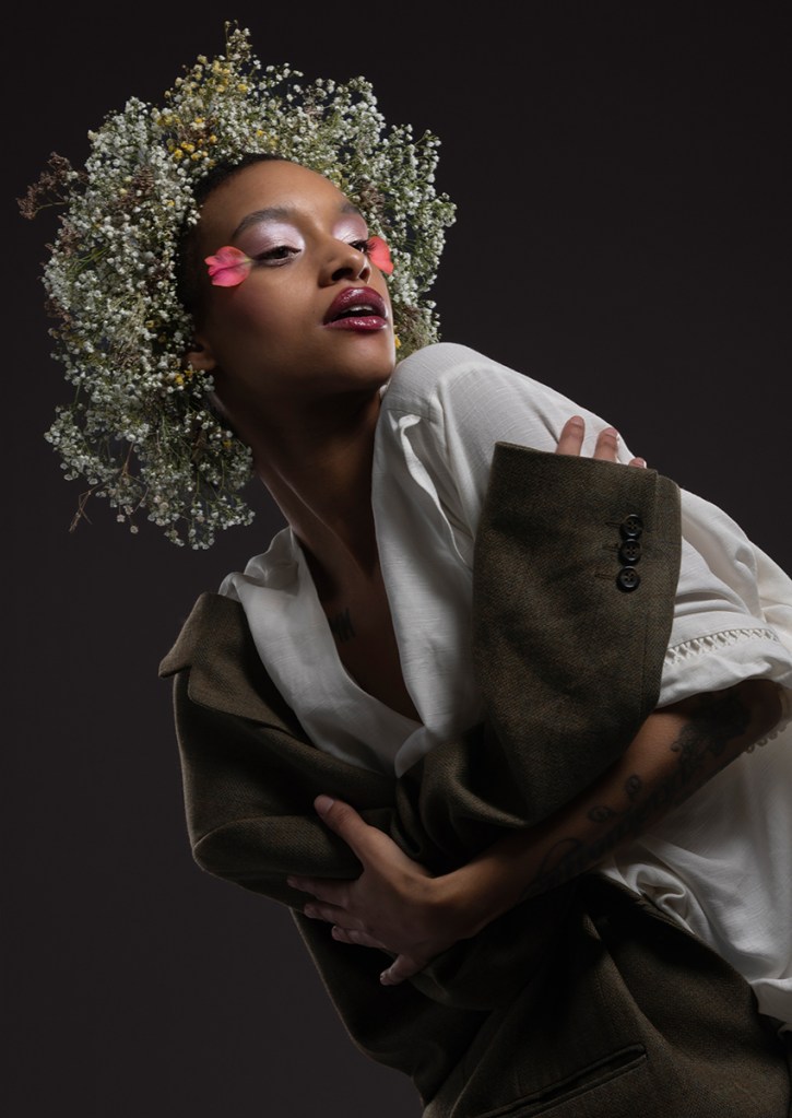 'Flower Crown' from Rooted in Her. Featuring a model in a green blazer and an intricate flower headpiece, this image expands on the themes of elegance and confidence born from a connection with the earth. It illustrates how women derive strength and authority from the communal support of gardeners, a network that directly elevates their well-being. Captured from a low angle to project height and stature, the portrait utilizes the model’s relaxed, creative styling of the blazer to project a quiet dominance. Ultimately, this confidence is rooted in a nurturing power, highlighting the profound authority that comes from knowing how to care for and appreciate nature. I