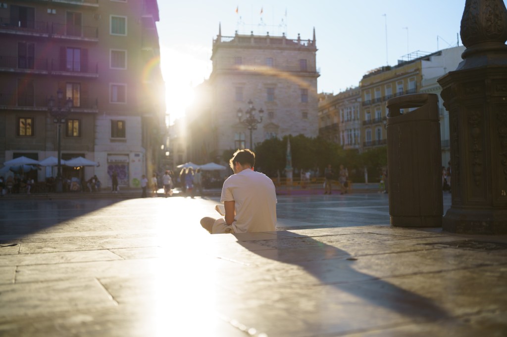 Viltrox AF 35mm F1.8 EVO II sample image. A man sitting on a town square with the sun peaking out from two buildings is illuminating him and casting a long shadow in the foreground