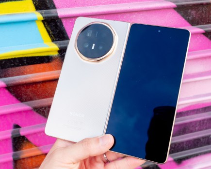 Honor Magic V6 camera phone in hand, unfolded, with its rear and outer screen showing. Photographed against a pink graffiti wall.
