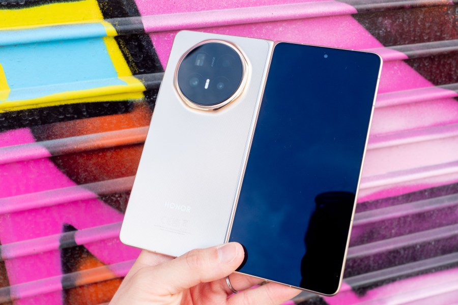 Honor Magic V6 camera phone in hand, unfolded, with its rear and outer screen showing. Photographed against a pink graffiti wall.