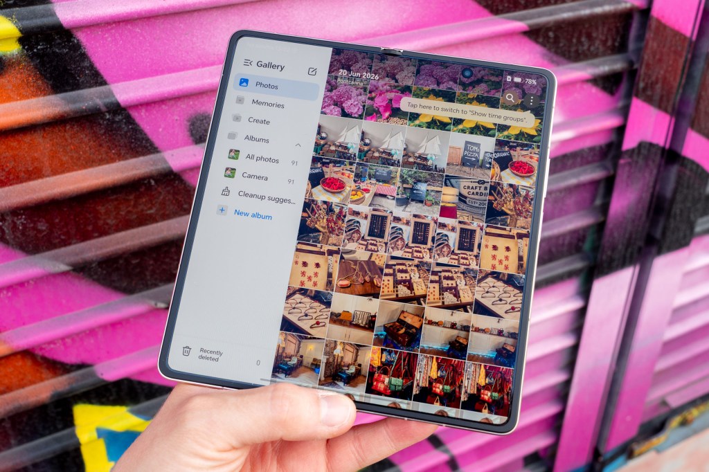 Honor Magic V6 foldable camera phone in hand displaying the gallery app on the large inner screen. Photographed against a pink graffiti wall